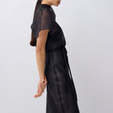 A woman wearing a black sheer short-sleeve top and matching black satin-like skirt with a tie belt, shown in profile against a light gray studio background.