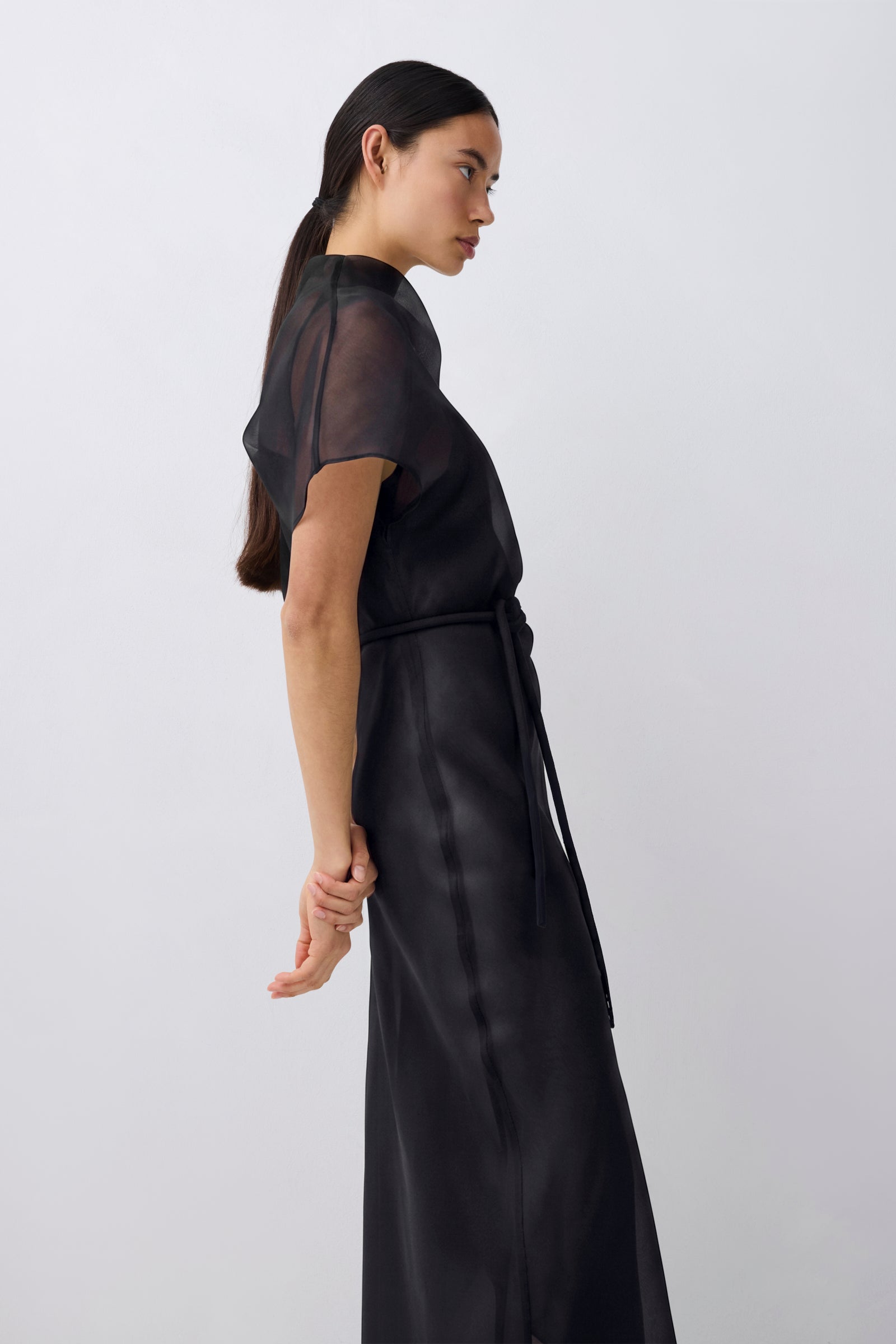 A woman wearing a black sheer short-sleeve top and matching black satin-like skirt with a tie belt, shown in profile against a light gray studio background.