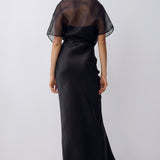 A woman viewed from the back wearing a long black dress with a sheer mesh short-sleeve overlay and a satin-like skirt, photographed against a plain light gray studio background.