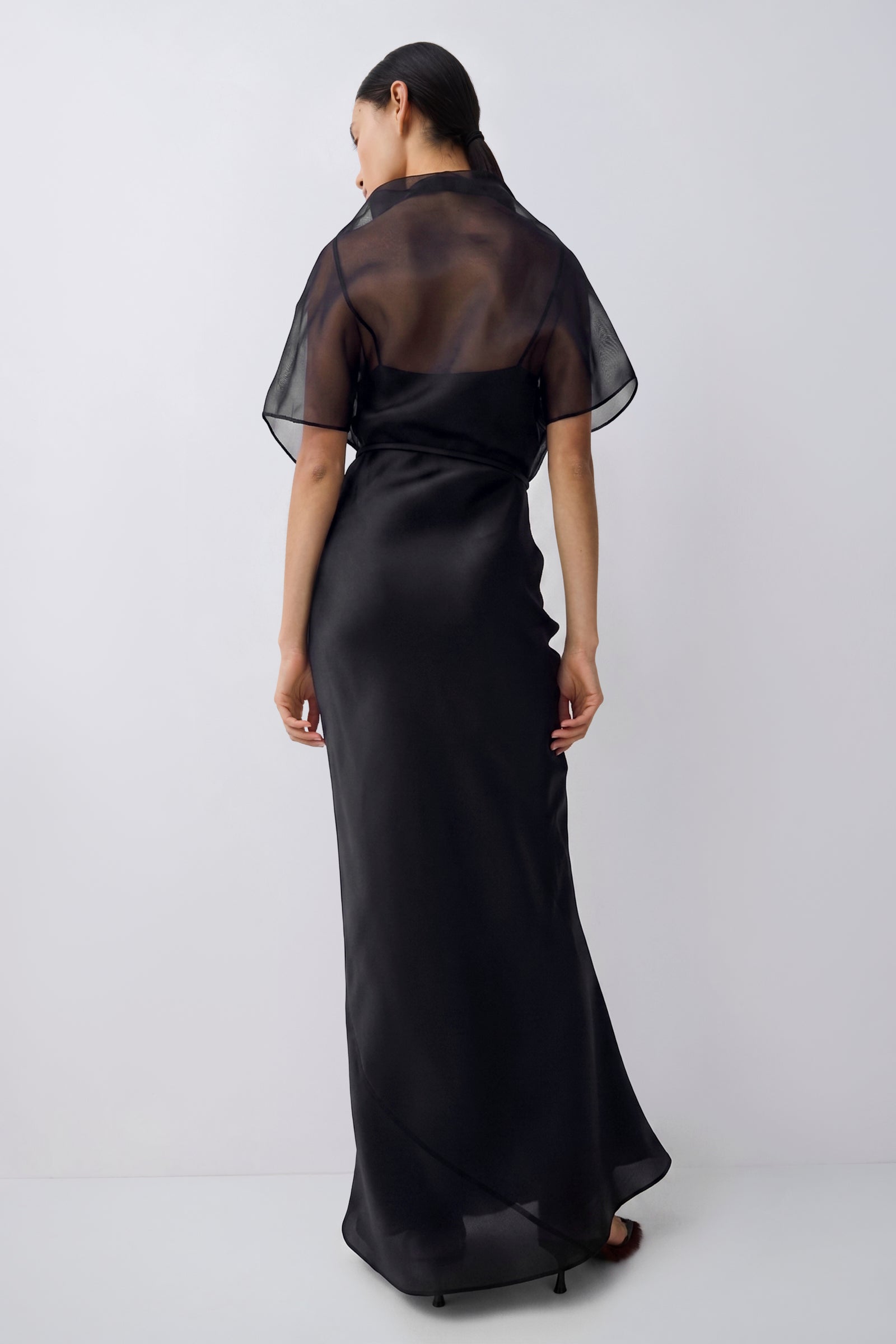 A woman viewed from the back wearing a long black dress with a sheer mesh short-sleeve overlay and a satin-like skirt, photographed against a plain light gray studio background.