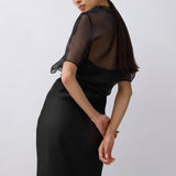 A woman modeling a black midi dress with sheer short sleeves and a draped open back, photographed against a plain light gray studio background.