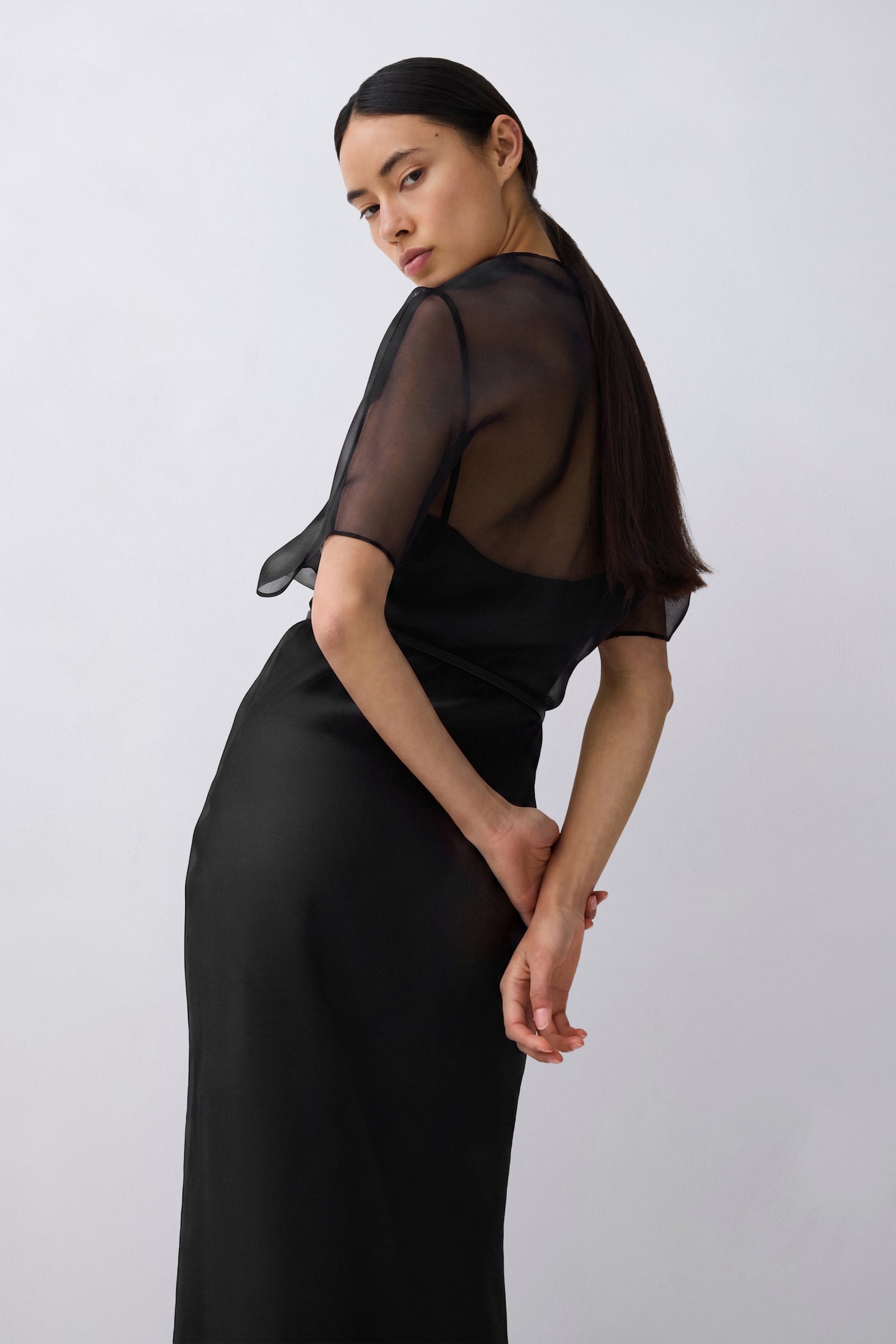 A woman modeling a black midi dress with sheer short sleeves and a draped open back, photographed against a plain light gray studio background.