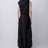 A woman viewed from behind wearing a sleeveless black maxi dress with a draped high neckline and a gathered skirt, standing against a light gray studio background.