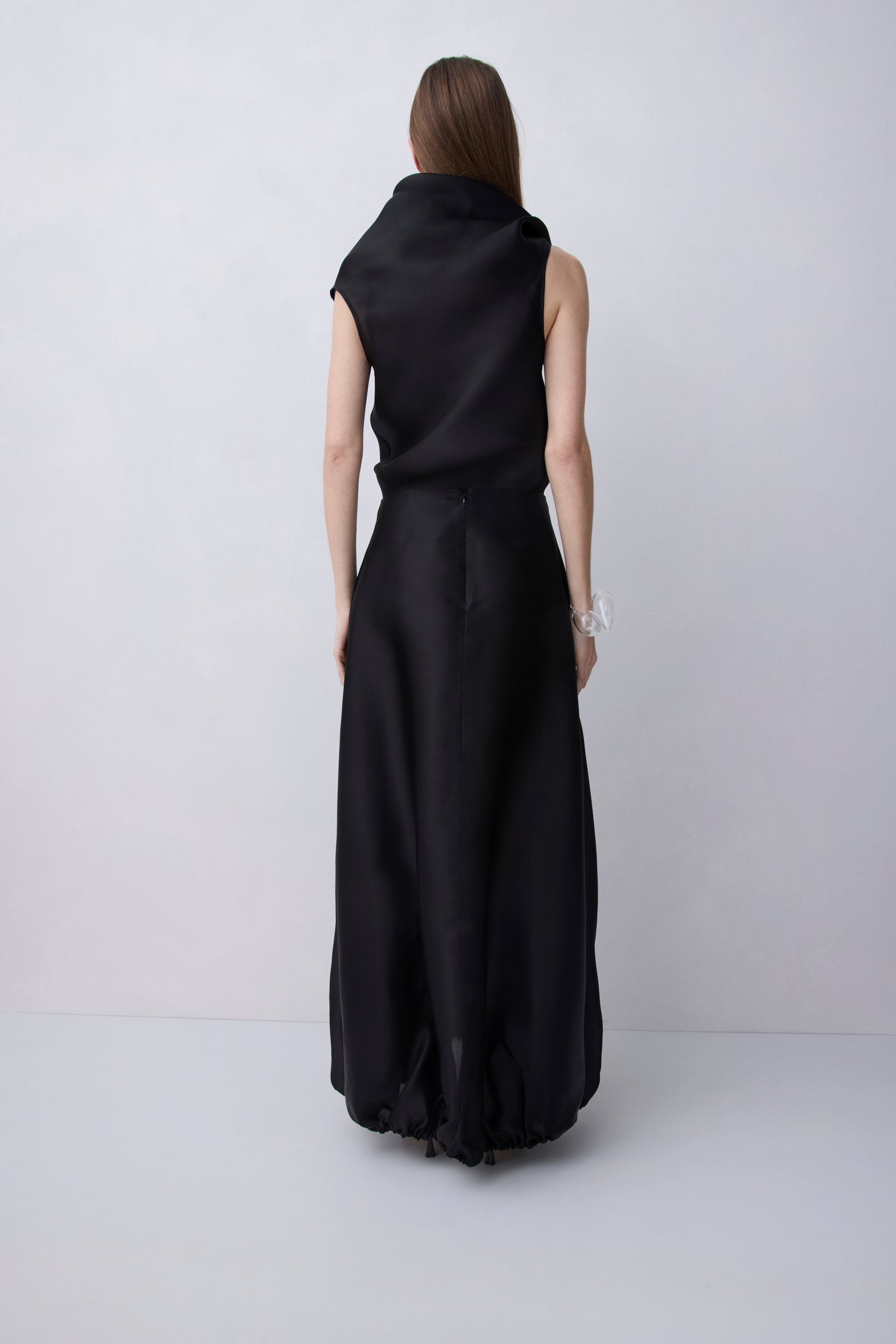 A woman viewed from behind wearing a sleeveless black maxi dress with a draped high neckline and a gathered skirt, standing against a light gray studio background.