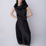 A woman modeling a black sleeveless maxi dress with a dramatic oversized draped neckline and voluminous gathered skirt, standing against a light gray studio background.