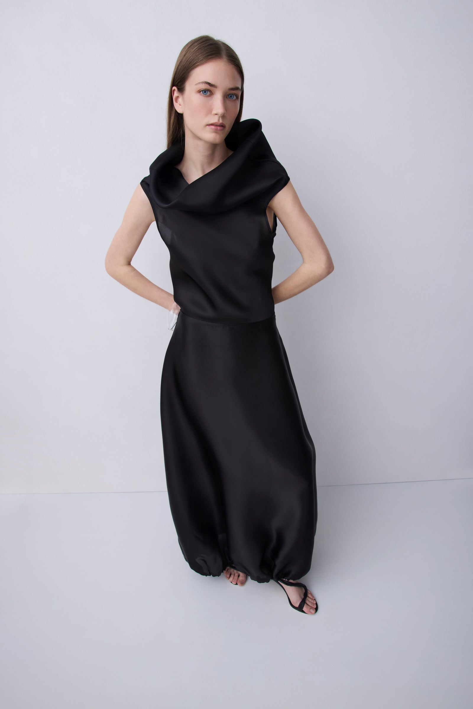 A woman modeling a black sleeveless maxi dress with a dramatic oversized draped neckline and voluminous gathered skirt, standing against a light gray studio background.