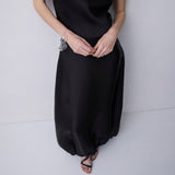 A model wearing a black satin-like cowl-neck maxi dress with a gathered, balloon-style skirt and black strappy flat sandals, standing against a light gray background.
