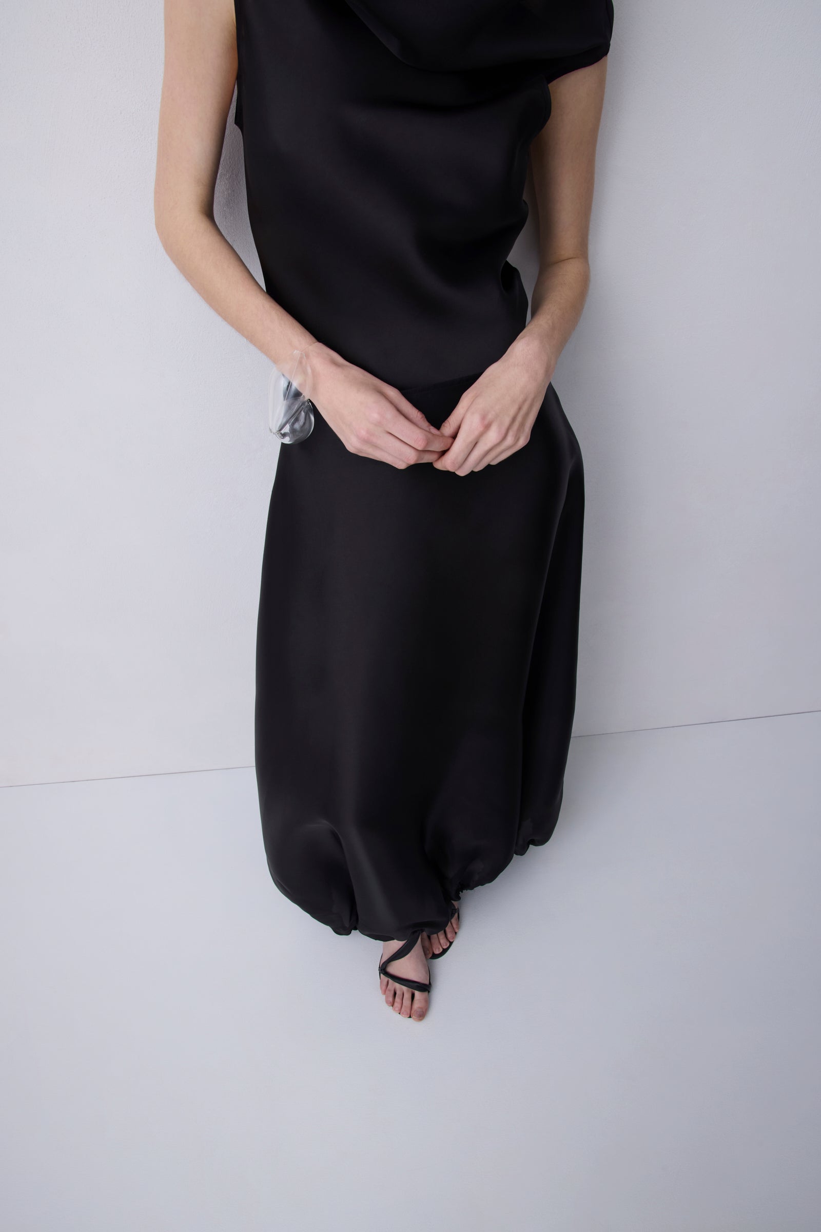 A model wearing a black satin-like cowl-neck maxi dress with a gathered, balloon-style skirt and black strappy flat sandals, standing against a light gray background.