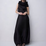 A woman modeling a floor-length black satin gown with a draped, asymmetrical neckline and voluminous skirt, standing against a light gray studio background.