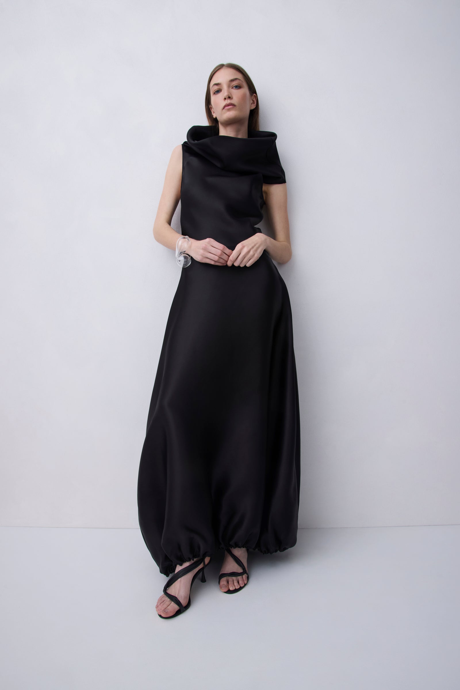 A woman modeling a floor-length black satin gown with a draped, asymmetrical neckline and voluminous skirt, standing against a light gray studio background.