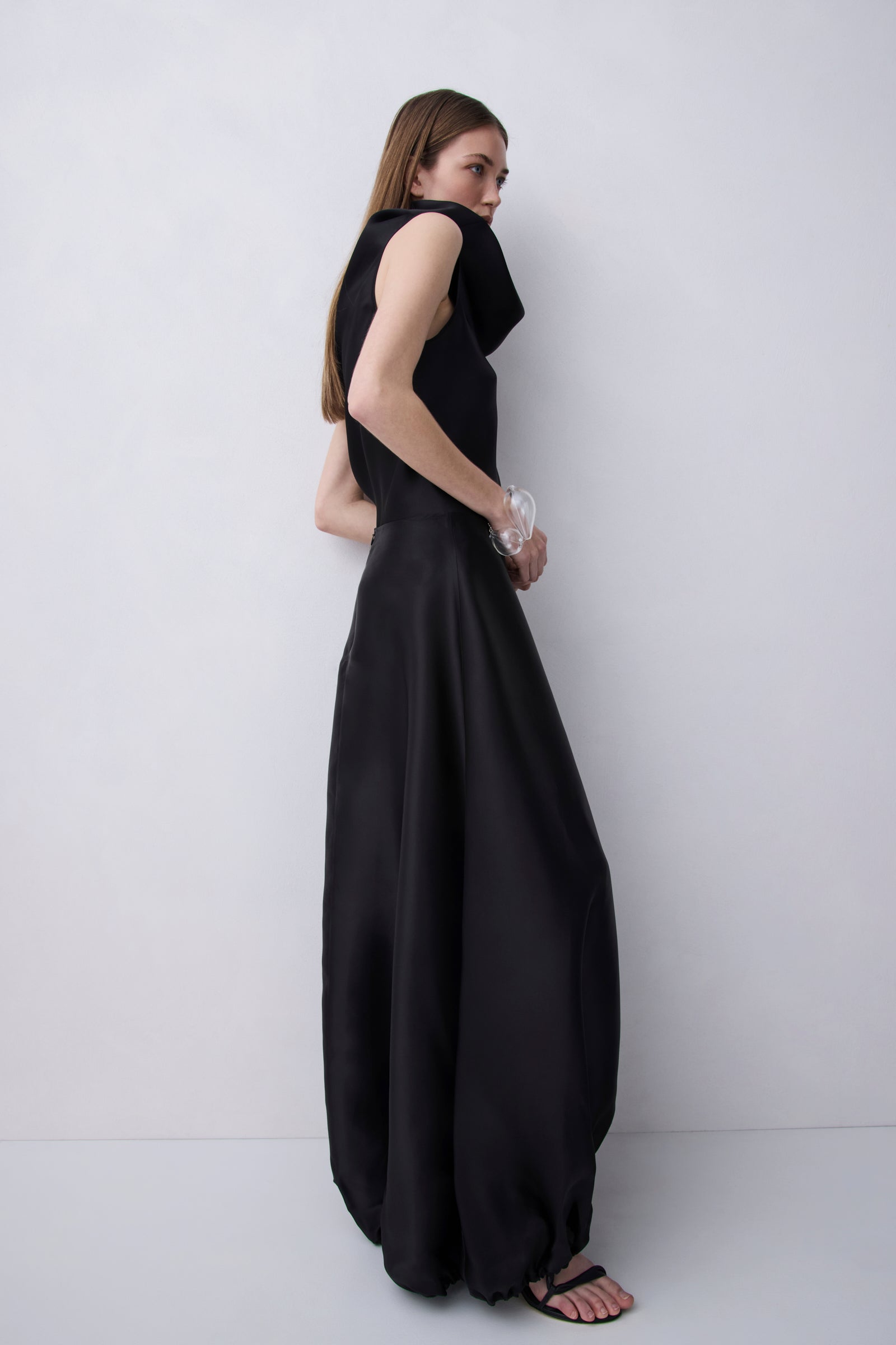 A woman modeling a sleeveless black maxi dress with a draped, voluminous skirt, photographed against a plain light gray studio background.