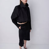 A model wearing a black cropped long-sleeve top with a high gathered collar and a matching black midi skirt, paired with black strappy sandals, standing against a light gray studio background.