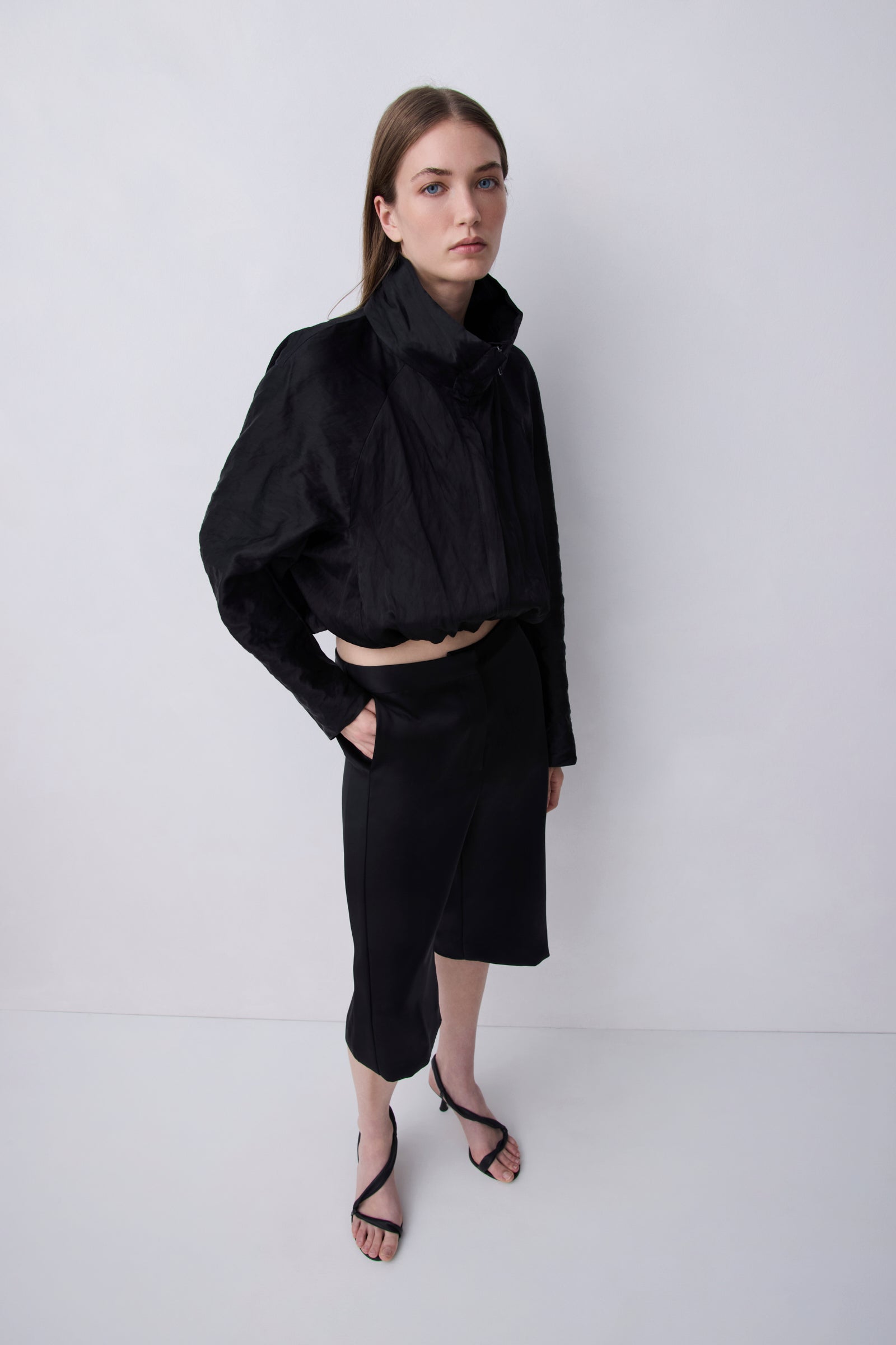 A model wearing a black cropped long-sleeve top with a high gathered collar and a matching black midi skirt, paired with black strappy sandals, standing against a light gray studio background.