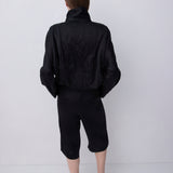 A model viewed from behind wearing a black cropped jacket with a high collar, black cropped pants, and black strappy heels, set against a plain light gray studio background.