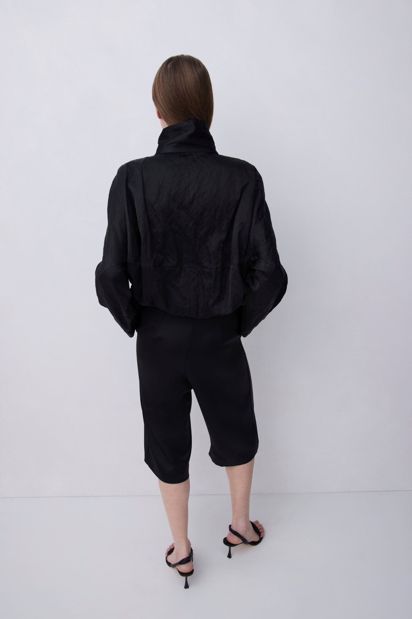 A model viewed from behind wearing a black cropped jacket with a high collar, black cropped pants, and black strappy heels, set against a plain light gray studio background.