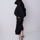 A model wearing a black high-neck, voluminous long-sleeve top and matching black cropped pants, paired with black strappy heels, standing against a plain light gray studio background.