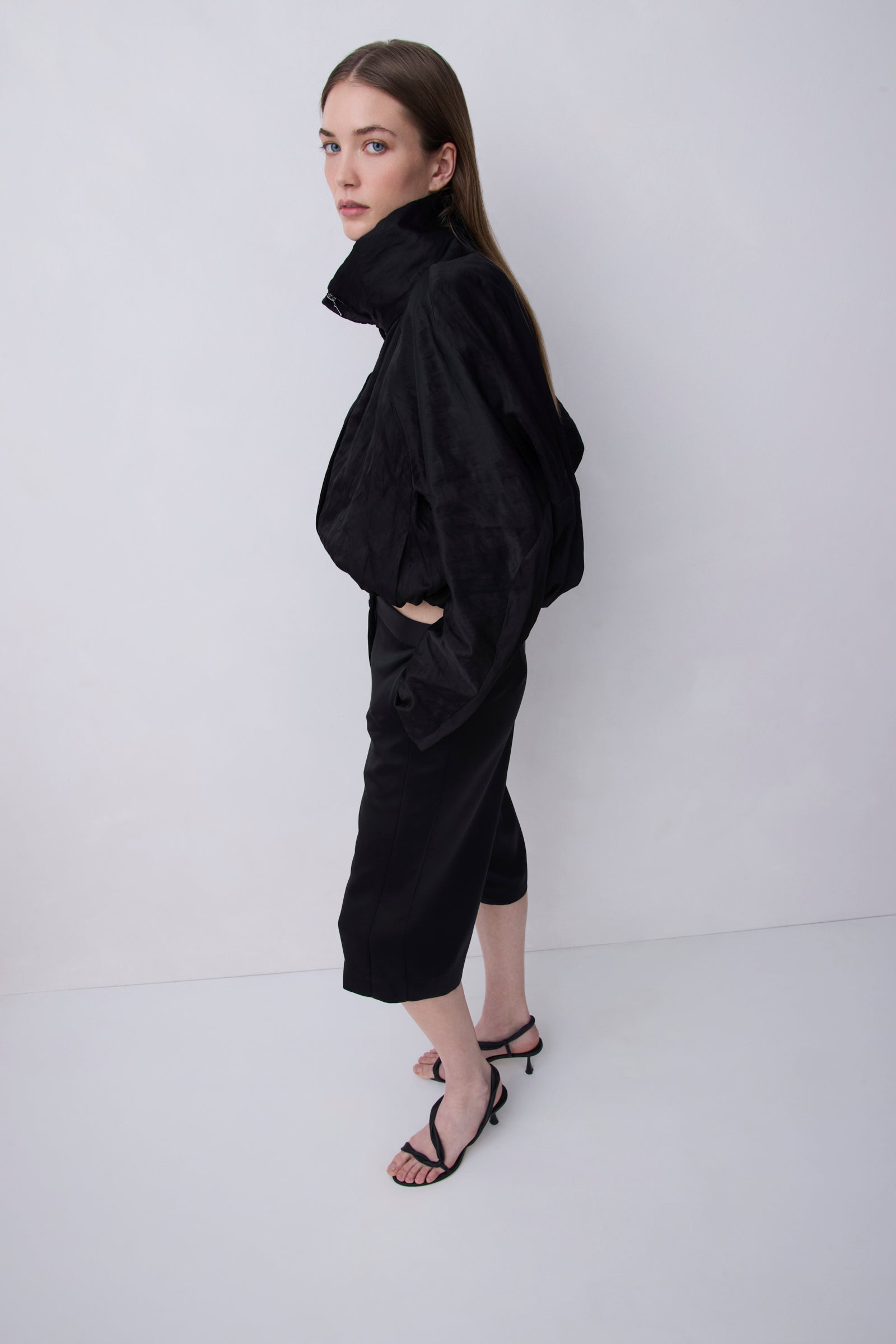 A model wearing a black high-neck, voluminous long-sleeve top and matching black cropped pants, paired with black strappy heels, standing against a plain light gray studio background.