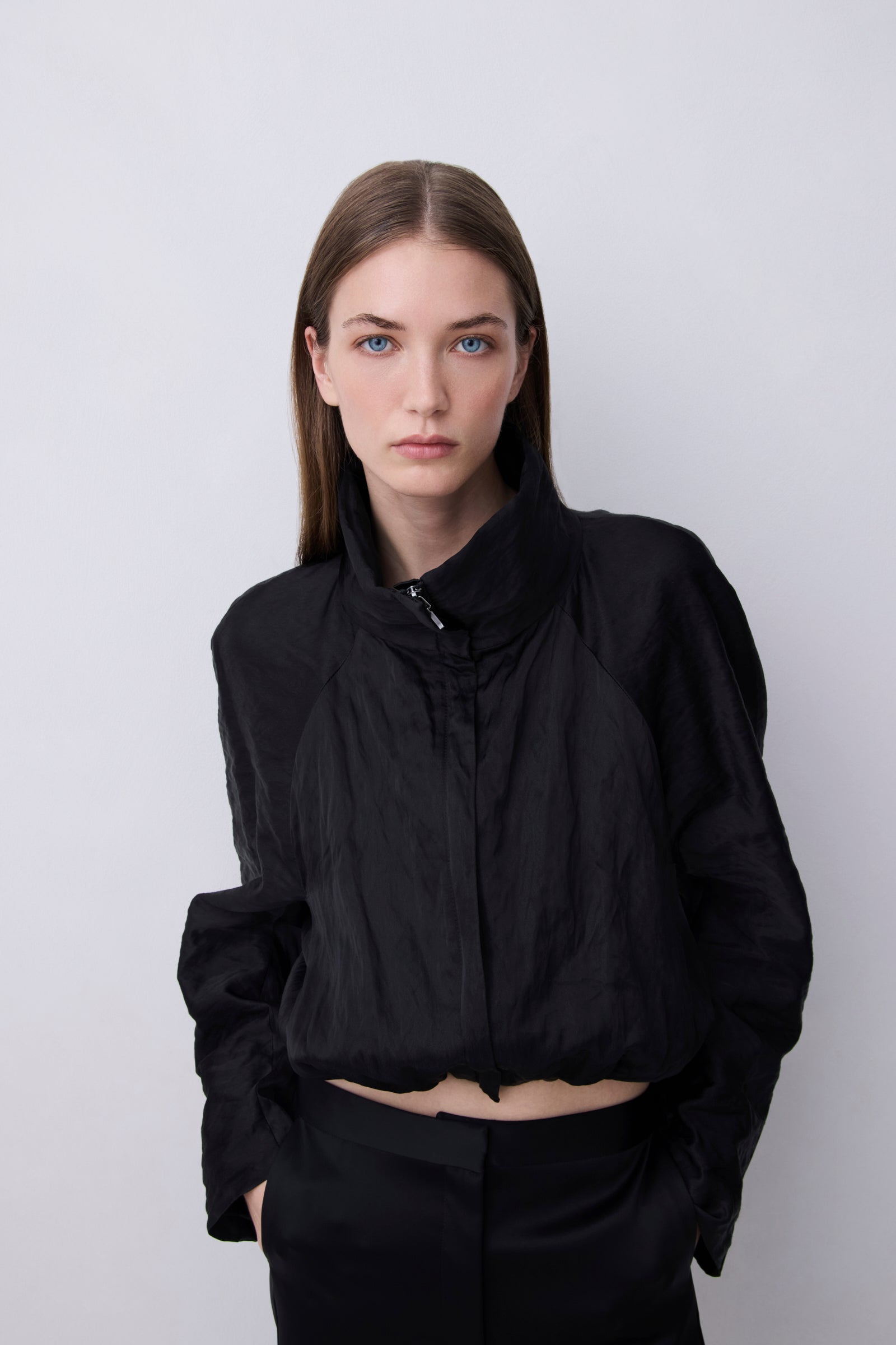A model wearing a black cropped lightweight jacket with a high collar and front zipper, paired with black pants, set against a plain light gray studio background.
