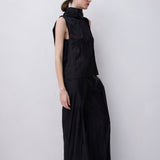 A model wearing a black sleeveless mock-neck top and matching wide-leg trousers, standing in profile against a plain light gray studio background.