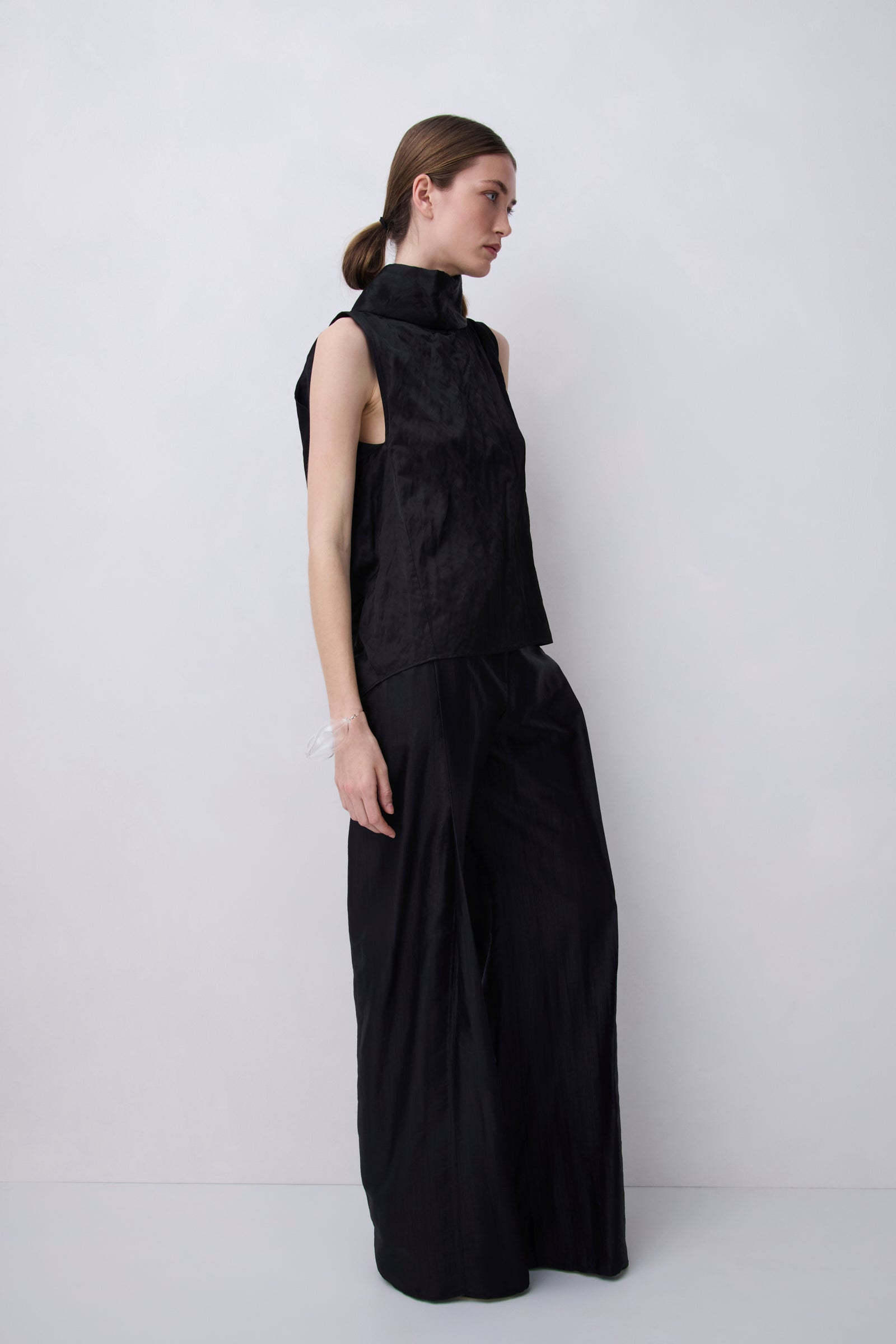 A model wearing a black sleeveless mock-neck top and matching wide-leg trousers, standing in profile against a plain light gray studio background.