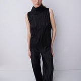 A model stands against a light gray studio background wearing a black sleeveless mock-neck top and matching wide-leg trousers.
