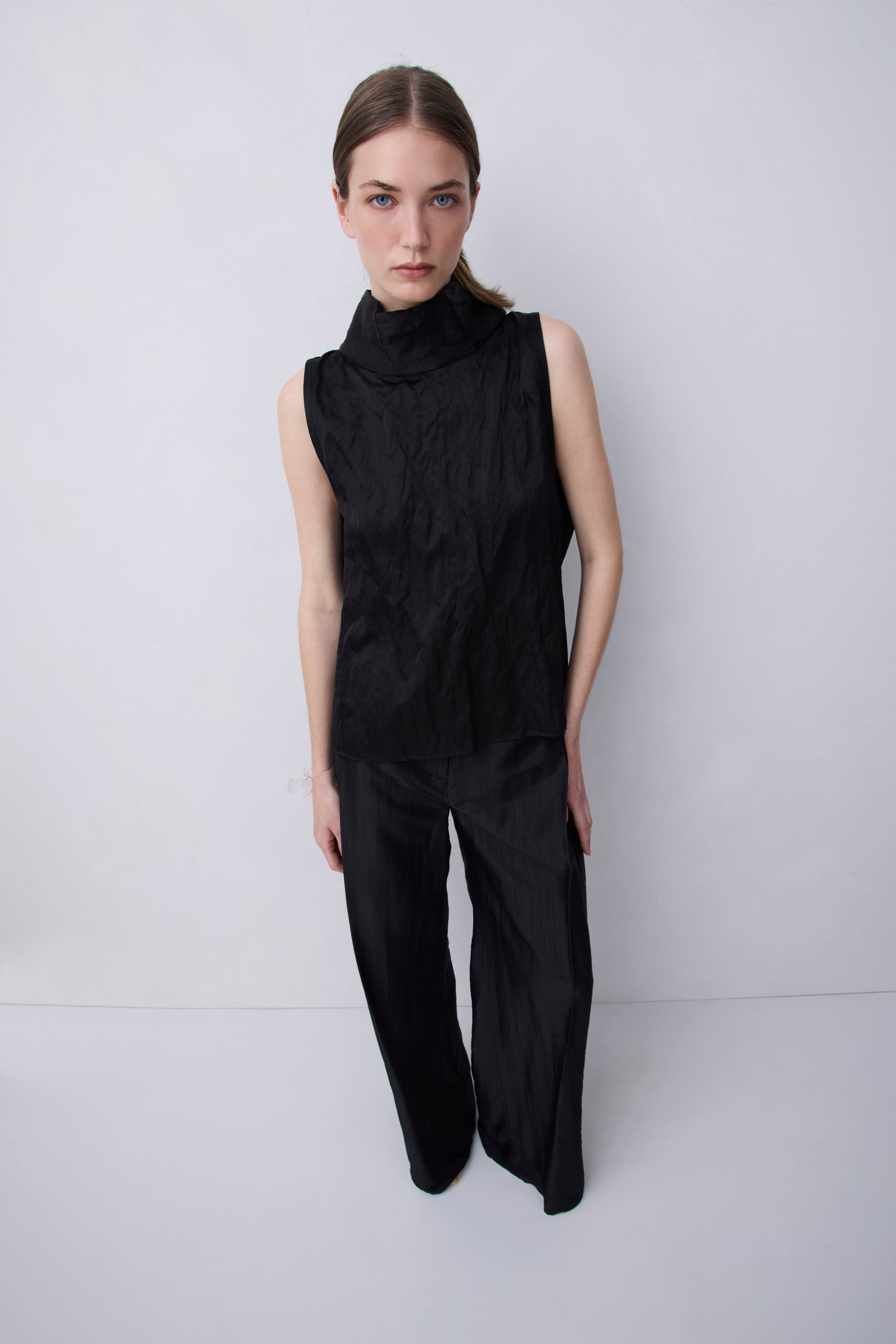 A model stands against a light gray studio background wearing a black sleeveless mock-neck top and matching wide-leg trousers.