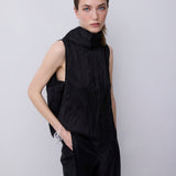 A model wearing a black sleeveless high-neck top and matching black wide-leg trousers, standing against a light gray studio background.