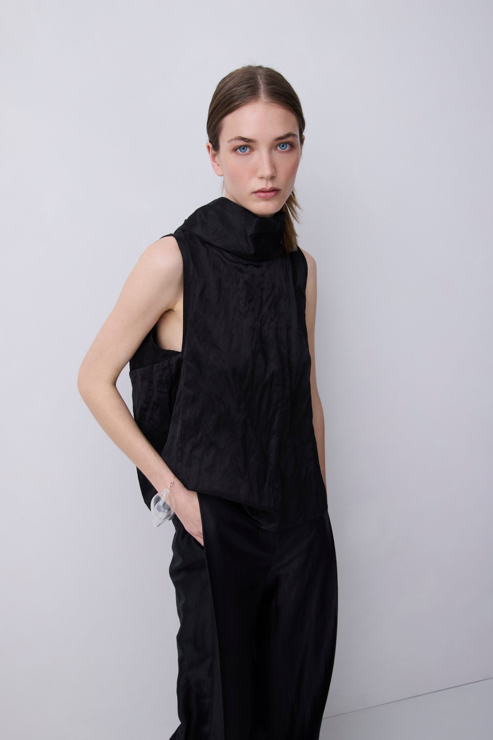 A model wearing a black sleeveless high-neck top and matching black wide-leg trousers, standing against a light gray studio background.