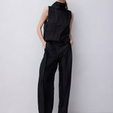 A model stands against a light gray studio background wearing a black sleeveless high-neck top and matching wide-leg trousers.