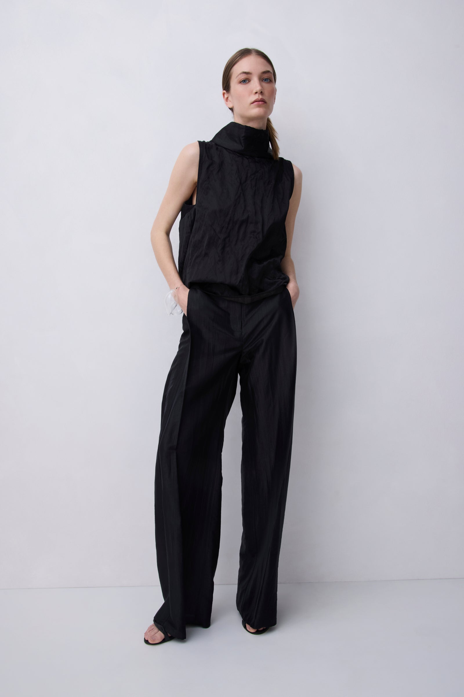 A model stands against a light gray studio background wearing a black sleeveless high-neck top and matching wide-leg trousers.