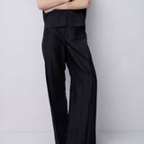 A model wearing a black sleeveless high-neck top and matching black wide-leg trousers, standing with arms crossed against a light gray studio background.