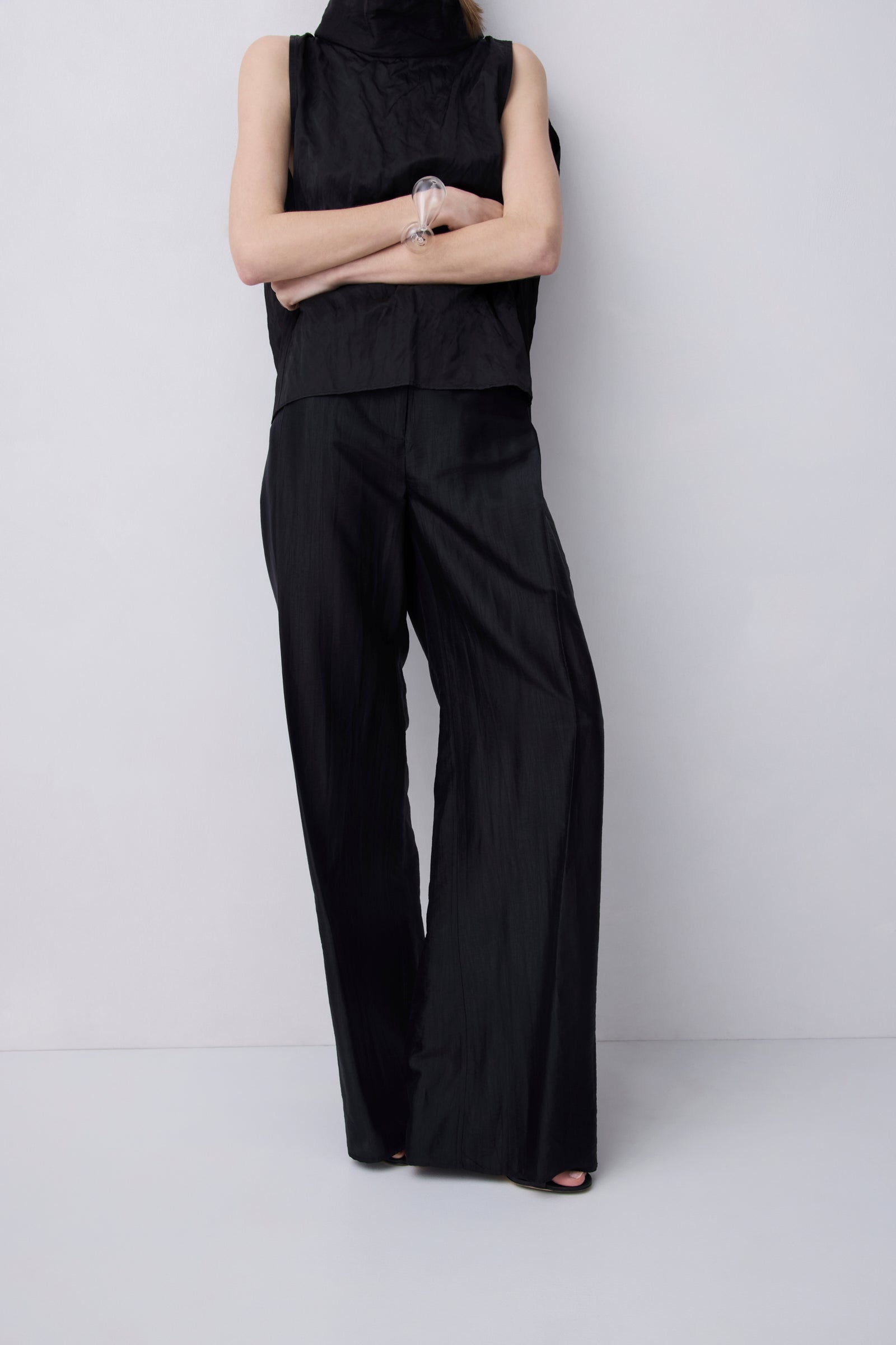 A model wearing a black sleeveless high-neck top and matching black wide-leg trousers, standing with arms crossed against a light gray studio background.