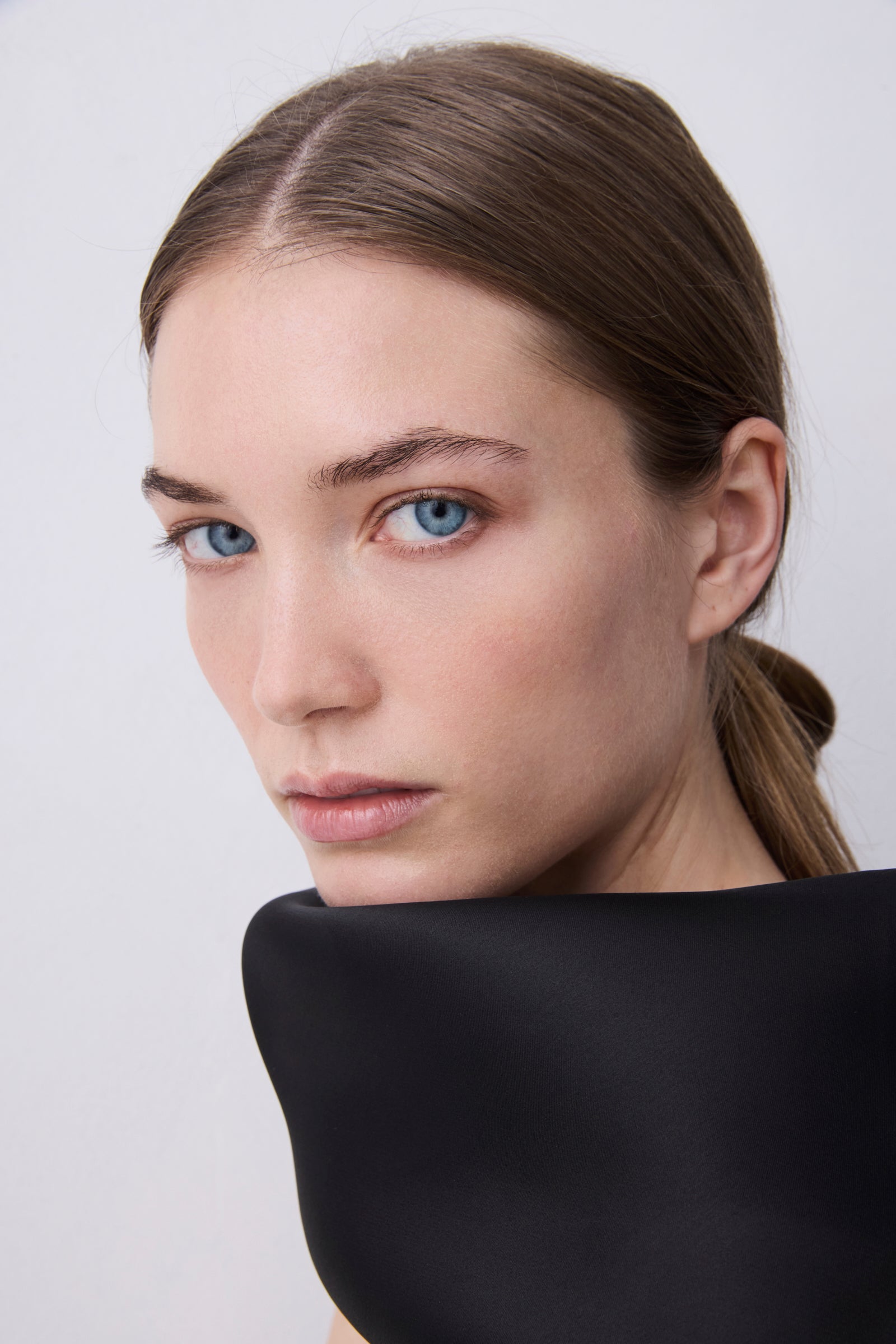 A close-up portrait of a woman with brown hair pulled back, wearing a black high-neck top against a light gray background.