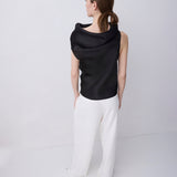 A woman shown from the back wearing a black draped sleeveless top and white wide-leg trousers, standing against a plain light gray studio background.