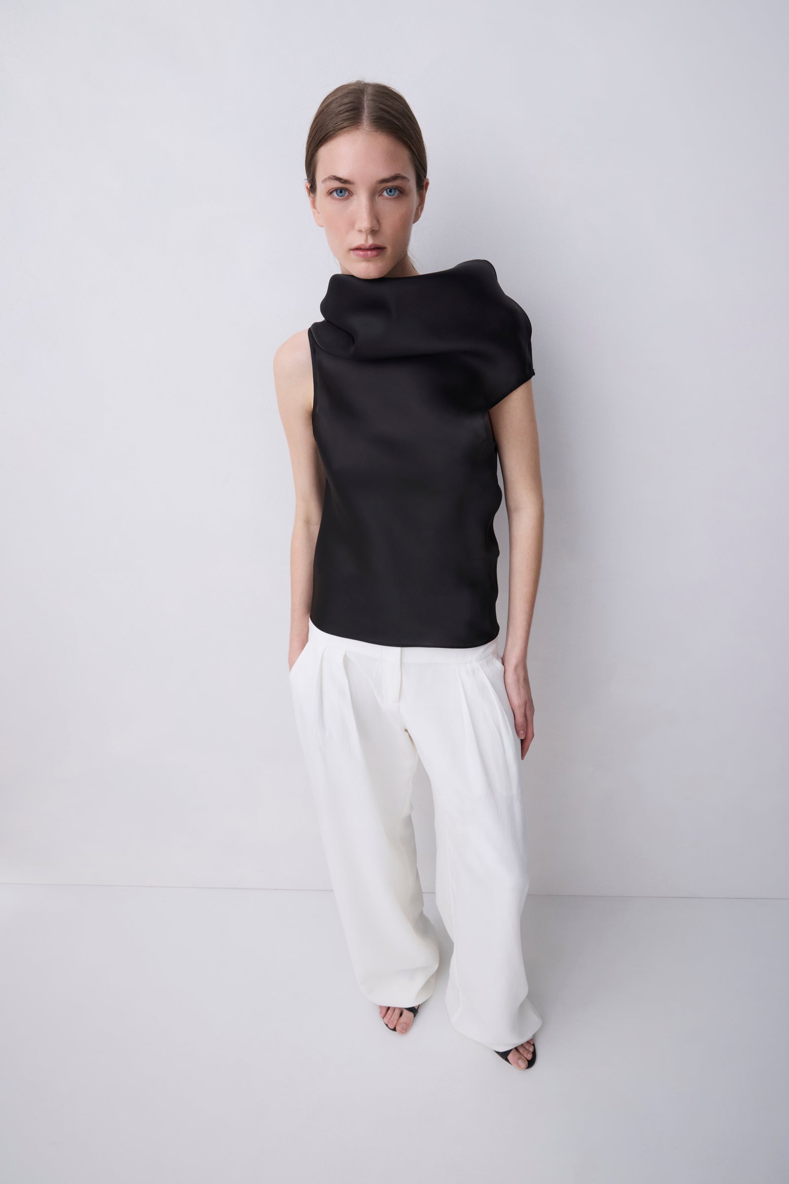 A model stands against a light gray studio background wearing a black draped sleeveless top and wide-leg white trousers.