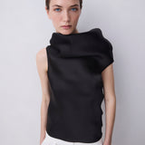 A model wearing a black draped, asymmetrical sleeveless top and white trousers, photographed against a neutral light gray background.