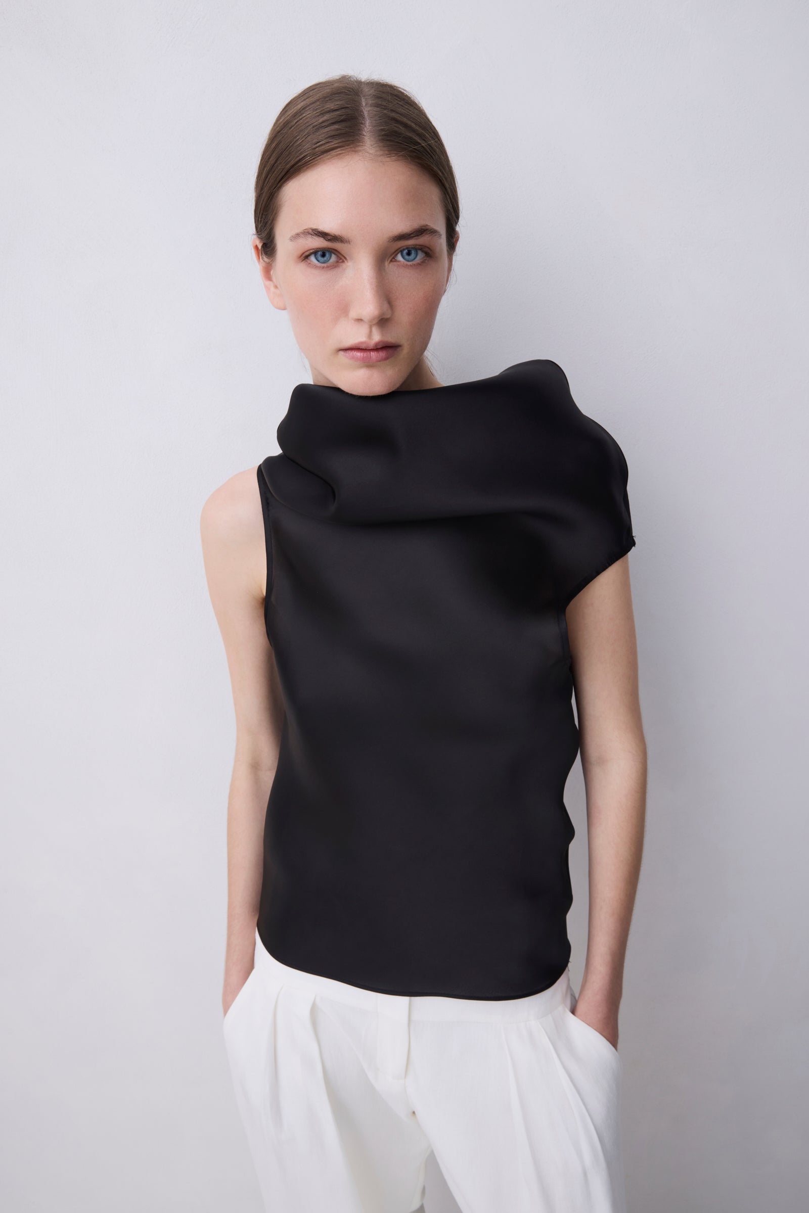 A model wearing a black draped, asymmetrical sleeveless top and white trousers, photographed against a neutral light gray background.