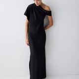 A woman modeling a floor-length black one-shoulder gown with a draped neckline and a side slit, standing against a plain light gray studio wall.