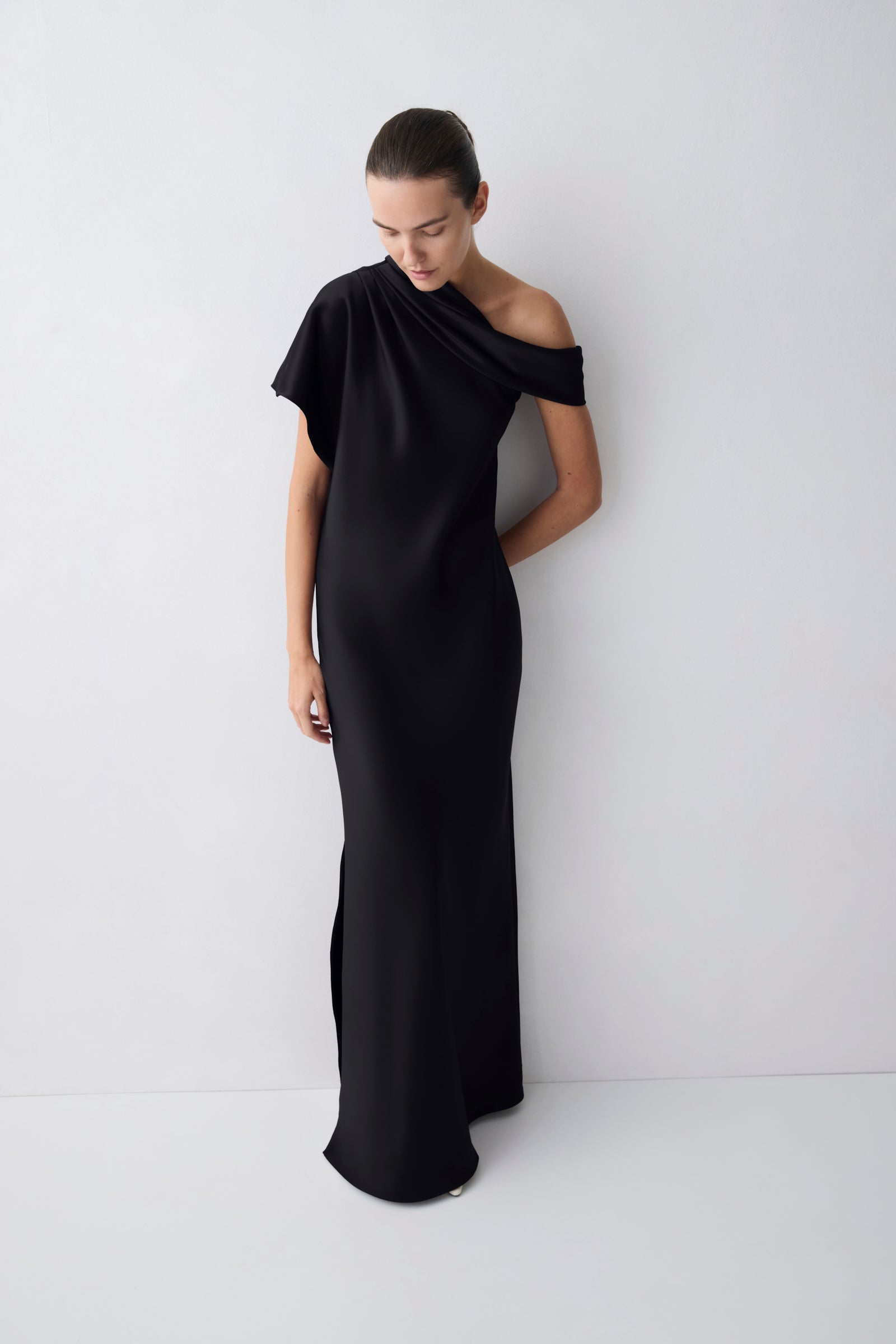 A woman modeling a floor-length black one-shoulder gown with a draped neckline and a side slit, standing against a plain light gray studio wall.