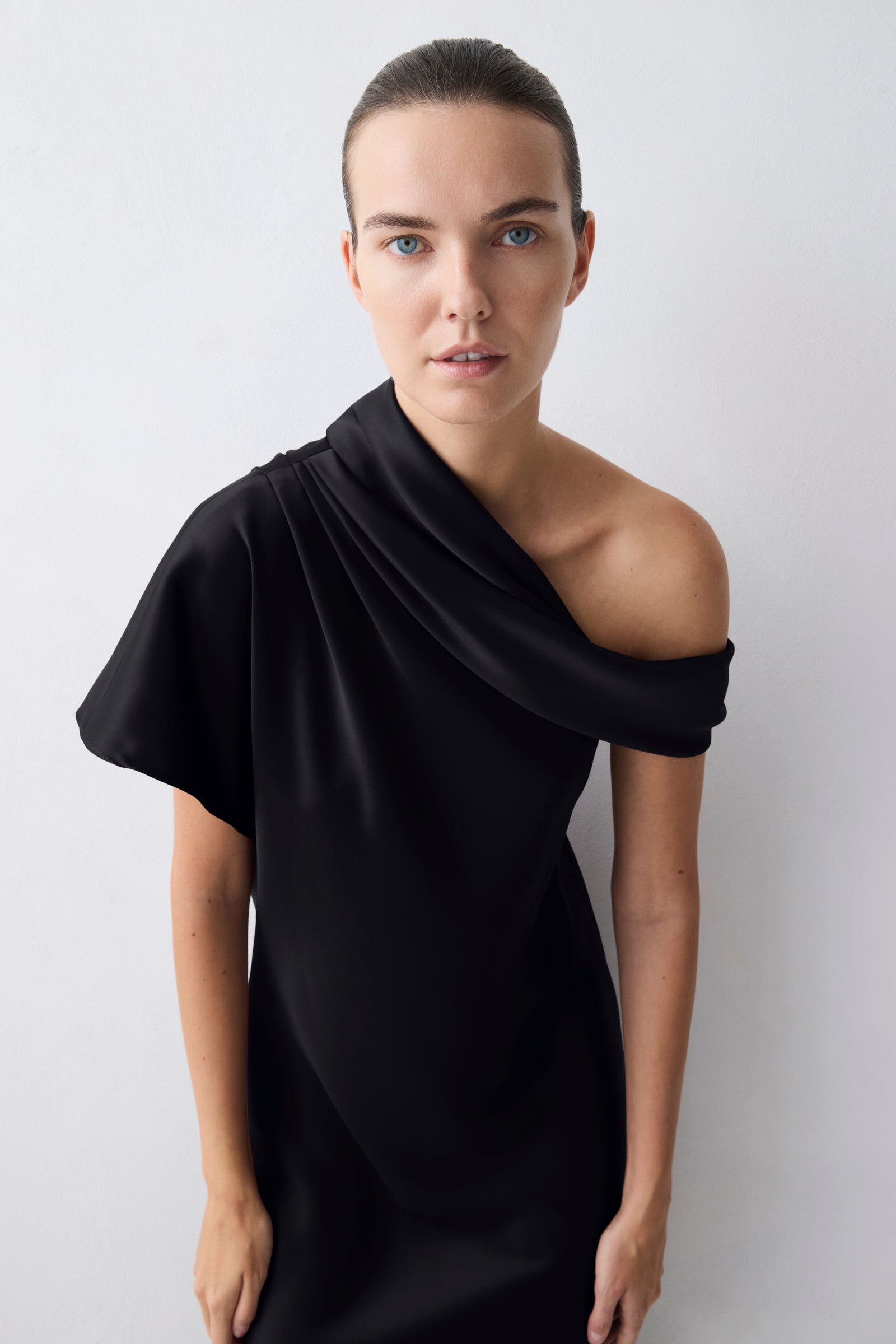 A model wearing a black draped one-shoulder dress against a light gray background.