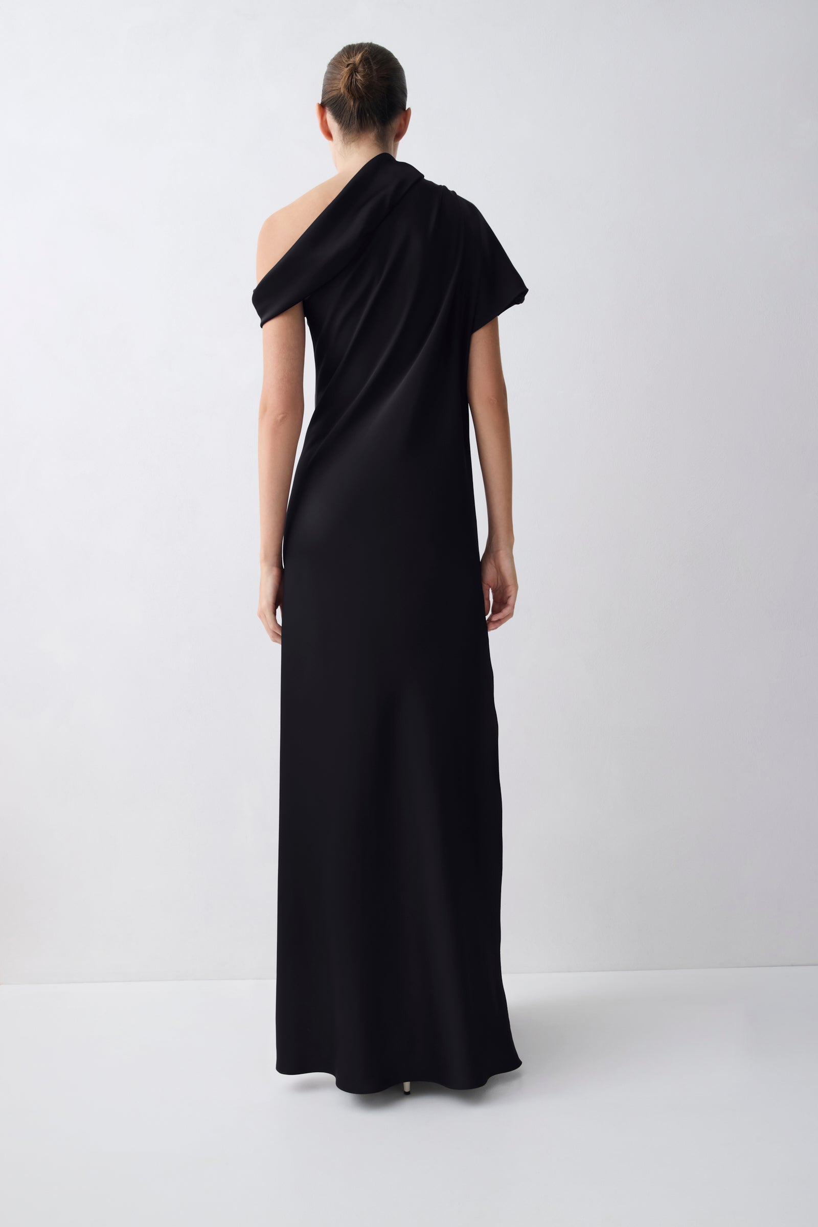 A model viewed from the back wearing a black one-shoulder maxi dress with short sleeves and a draped neckline, set against a plain light gray studio background.