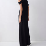 A woman wearing a long black off-the-shoulder maxi dress, photographed from the back in a minimalist studio setting with a light gray background.
