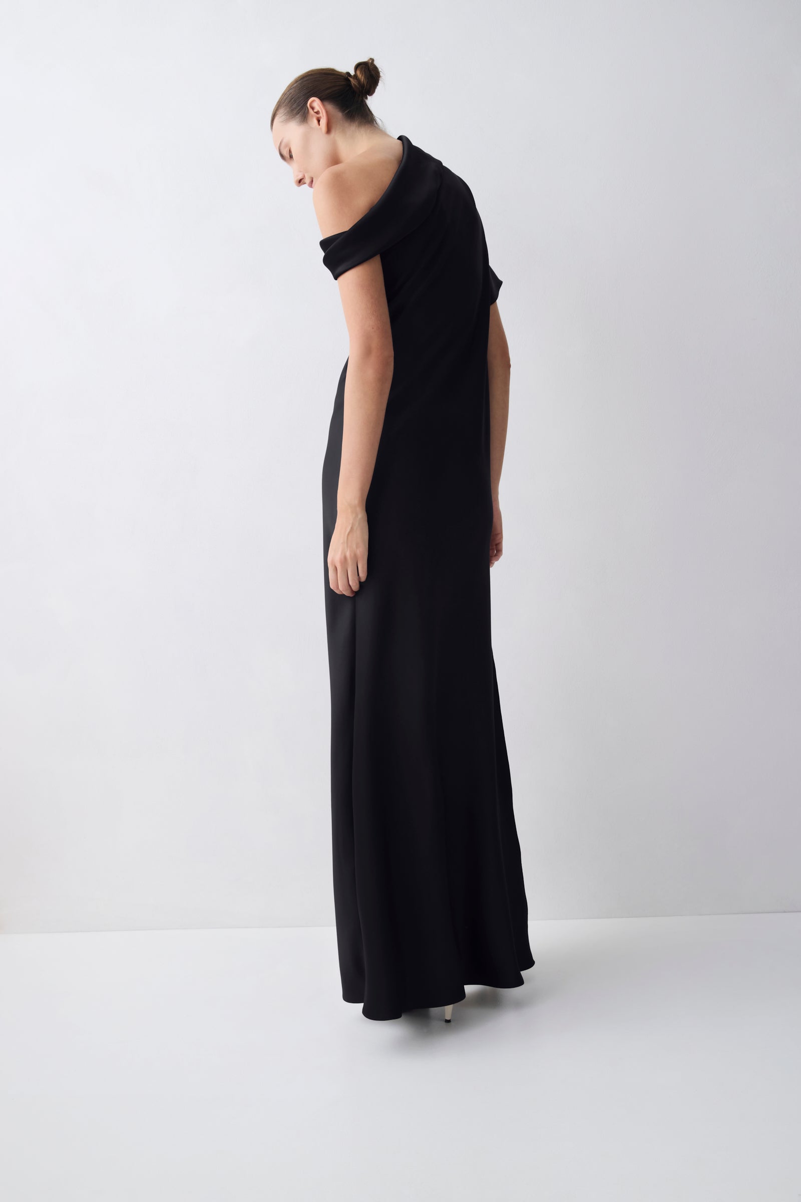 A woman wearing a long black off-the-shoulder maxi dress, photographed from the back in a minimalist studio setting with a light gray background.