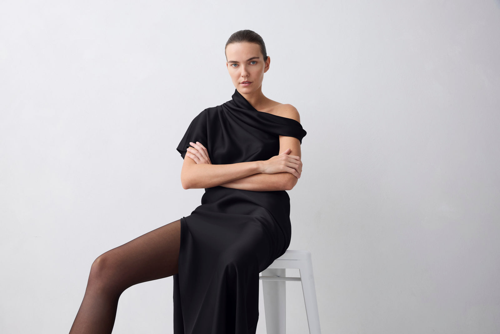 A woman seated on a white stool wearing a black draped off-the-shoulder dress and sheer black tights, posed against a plain light gray studio background.