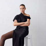 A woman seated on a white stool wearing a black draped off-the-shoulder dress and sheer black tights, posed against a plain light gray studio background.