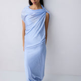 A model wearing a light blue draped maxi dress with short sleeves and a tie-neck detail, standing against a plain white studio background.