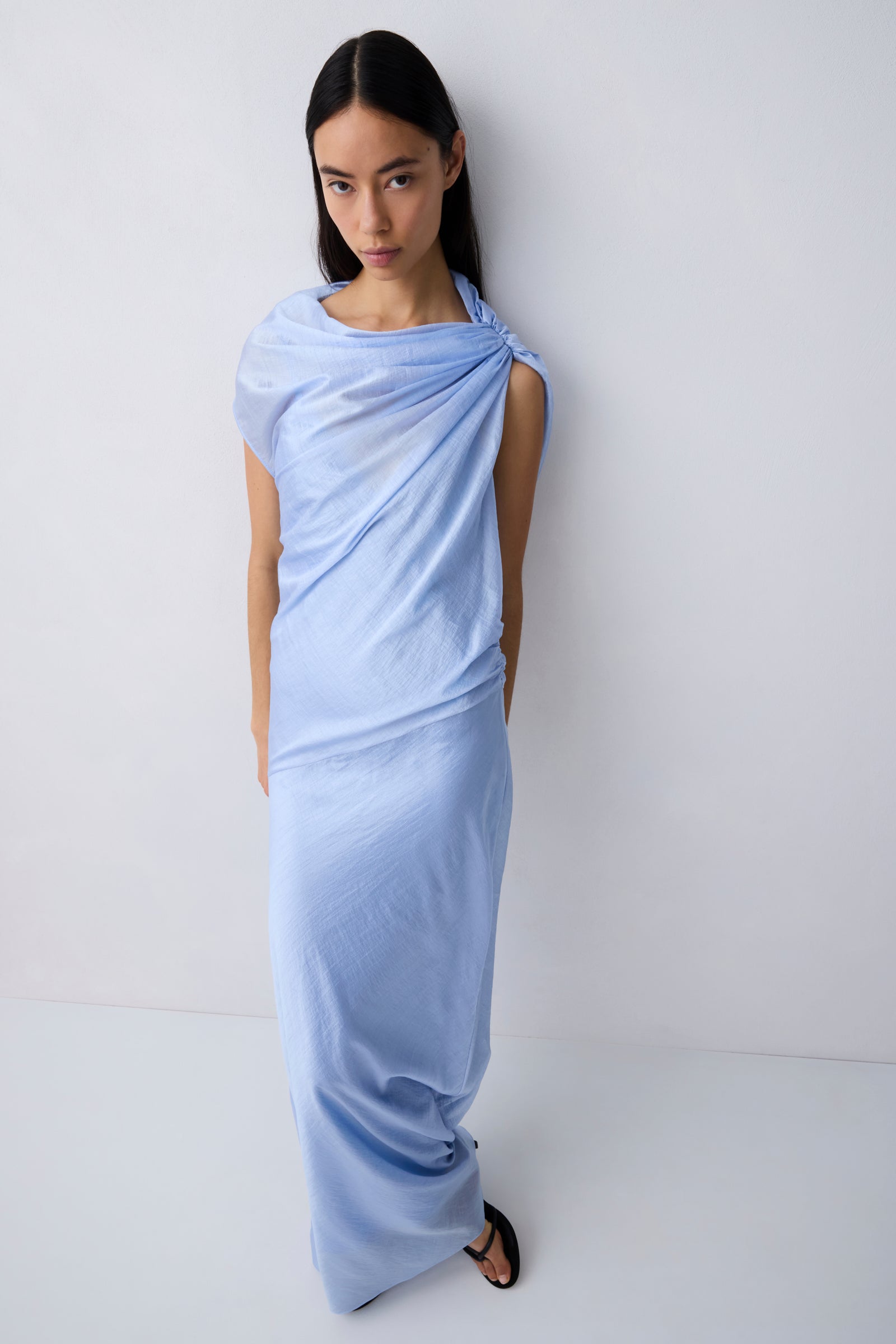 A model wearing a light blue draped maxi dress with short sleeves and a tie-neck detail, standing against a plain white studio background.