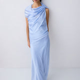 A model wearing a light blue draped maxi dress with short sleeves and black flat sandals, standing against a plain light gray studio background.