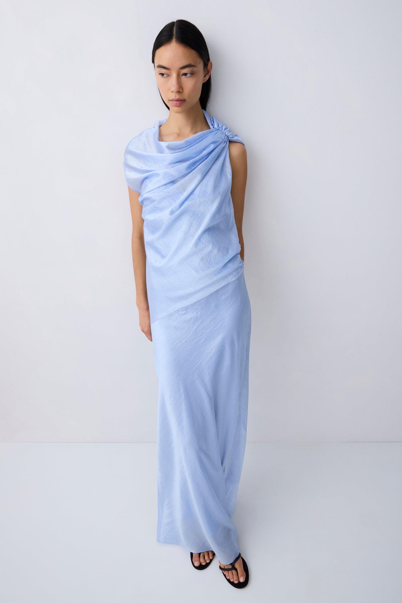 A model wearing a light blue draped maxi dress with short sleeves and black flat sandals, standing against a plain light gray studio background.