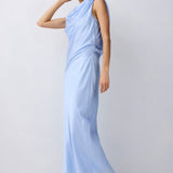 A woman modeling a light blue draped sleeveless maxi dress with a high neckline, standing against a plain light gray studio background.